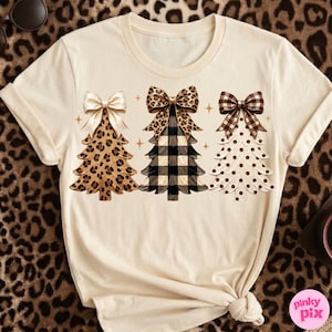 May include: A cream-colored t-shirt featuring a graphic of three stylized Christmas trees. The first tree has a leopard print pattern with a solid cream bow, the second has a black and white buffalo plaid pattern with a leopard print bow, and the third has a white polka dot pattern with a brown plaid bow.