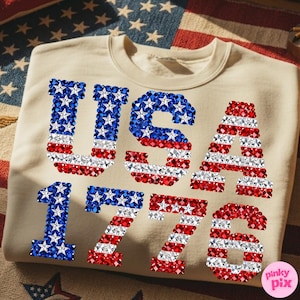 K&ouml;nnte beinhalten: Beiges Sweatshirt mit dem Text "USA 1776" in roten, wei&szlig;en und blauen Strasssteinen. Die Buchstaben und Zahlen sind im Design der amerikanischen Flagge mit Sternen und Streifen gehalten.