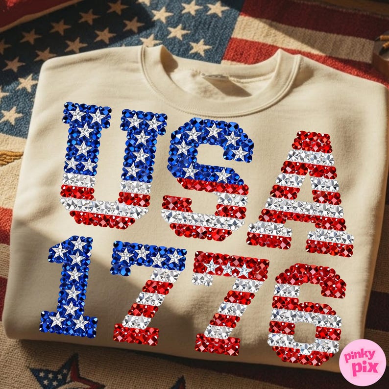 K&ouml;nnte beinhalten: Beiges Sweatshirt mit dem Text "USA 1776" in roten, wei&szlig;en und blauen Strasssteinen. Die Buchstaben und Zahlen sind im Design der amerikanischen Flagge mit Sternen und Streifen gehalten.
