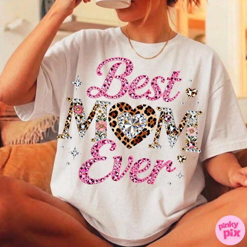 May include: White t-shirt with the text "Best Mom Ever" in pink, leopard print, and floral designs. The word "Mom" features a leopard print heart with a diamond-like center. The shirt has a crew neck and short sleeves.