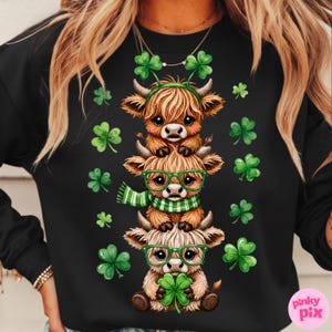 May include: Black sweatshirt featuring a stacked design of three cartoon Highland cows. The cows are brown and tan, wearing green glasses, scarves, and shamrock headbands. Green shamrocks are scattered around the cows.