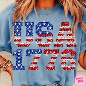 K&ouml;nnte beinhalten: Hellblaues T-Shirt mit dem Aufdruck "USA 1776". Die Buchstaben und Zahlen sind mit roten, wei&szlig;en und blauen Kristallen verziert. Ein l&auml;ssiges Oberteil mit patriotischem Thema, perfekt f&uuml;r Feiertage.
