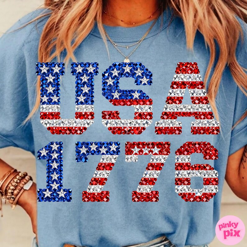 K&ouml;nnte beinhalten: Hellblaues T-Shirt mit dem Aufdruck "USA 1776". Die Buchstaben und Zahlen sind mit roten, wei&szlig;en und blauen Kristallen verziert. Ein l&auml;ssiges Oberteil mit patriotischem Thema, perfekt f&uuml;r Feiertage.