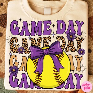 May include: Cream-colored sweatshirt with the words "GAME DAY" in purple, leopard print, and tan lettering. A yellow softball with a purple bow is centered on the design. Purple flowers and stars accent the design.