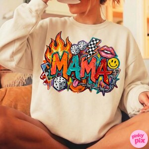 May include: Cream-colored sweatshirt featuring a vibrant graphic design. The design includes the word "MAMA" in bold, colorful letters, surrounded by flames, dice, a lightning bolt, lips, a smiley face, and barbed wire.