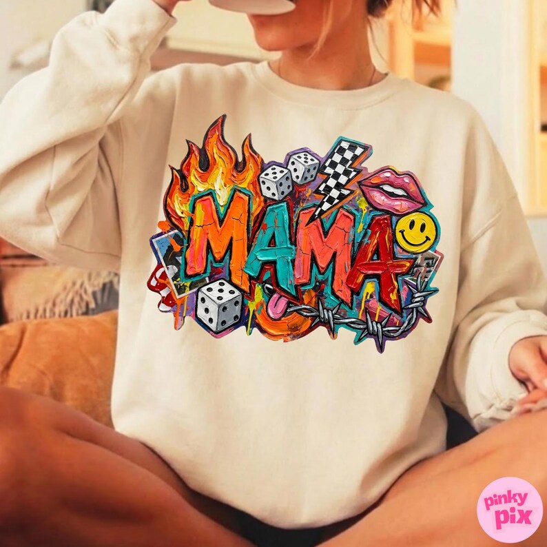 May include: Cream-colored sweatshirt featuring a vibrant graphic design. The design includes the word "MAMA" in bold, colorful letters, surrounded by flames, dice, a lightning bolt, lips, a smiley face, and barbed wire.