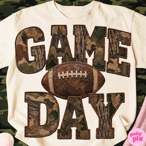 Könnte beinhalten: Ein cremefarbenes T-Shirt mit dem Schriftzug GAME DAY in großen Blockbuchstaben mit Tarn- und Baumrindenmuster. Zwischen den Wörtern befindet sich ein brauner Football mit weißen Nähten, der ebenfalls ein strukturiertes Muster aufweist.