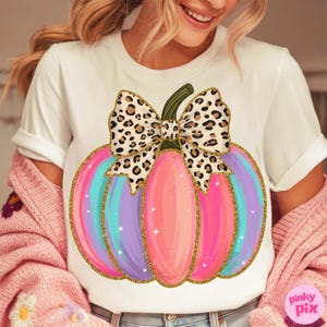 Puede incluir: Camiseta blanca con un diseño de calabaza colorida. La calabaza tiene rayas verticales en tonos rosa, morado y turquesa, delineadas con purpurina dorada. Un lazo con estampado de leopardo está encima. Es una prenda casual.