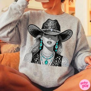 May include: A gray sweatshirt featuring a detailed graphic of a woman wearing a bedazzled cowboy hat, earrings, and necklace. The design is primarily black, white, and silver, with turquoise accents. The image has a rhinestone effect.