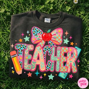 May include: Black sweatshirt with a colorful "TEACHER" design. The design includes a bow with a red apple, stars, a pencil, and a ruler. The letters are in various patterns and colors, including teal, orange, and pink.