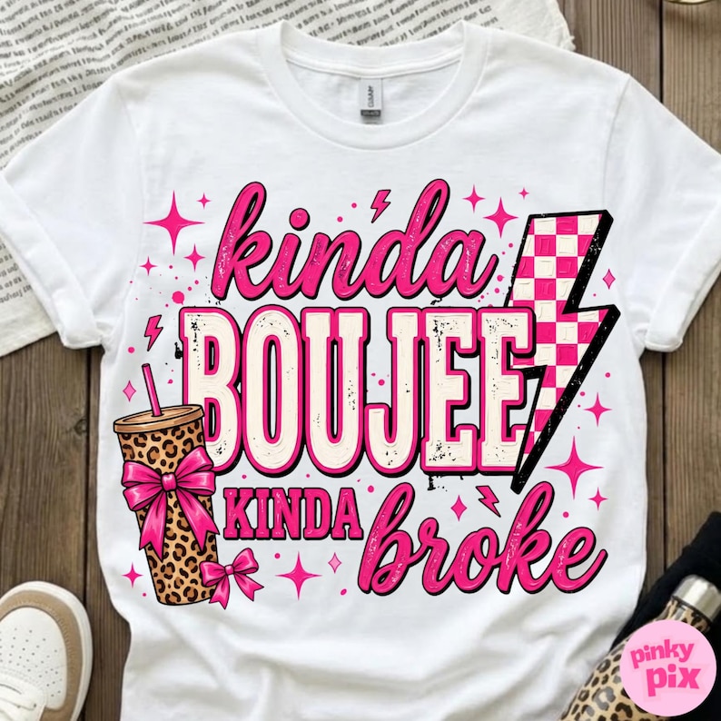 May include: A white short-sleeved t-shirt featuring a graphic print with the text "kinda BOUJEE kinda broke". The design includes a leopard print tumbler with a pink bow, a pink and white checkered lightning bolt, and pink star accents.