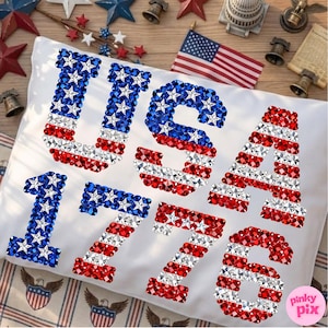 K&ouml;nnte beinhalten: Wei&szlig;es Kissen mit den W&ouml;rtern "USA" und den Zahlen "1776" in rot, wei&szlig; und blau glitzernden Verzierungen, die an die amerikanische Flagge erinnern. Das Kissen liegt auf einer Holzoberfl&auml;che mit patriotischen Dekorationen.