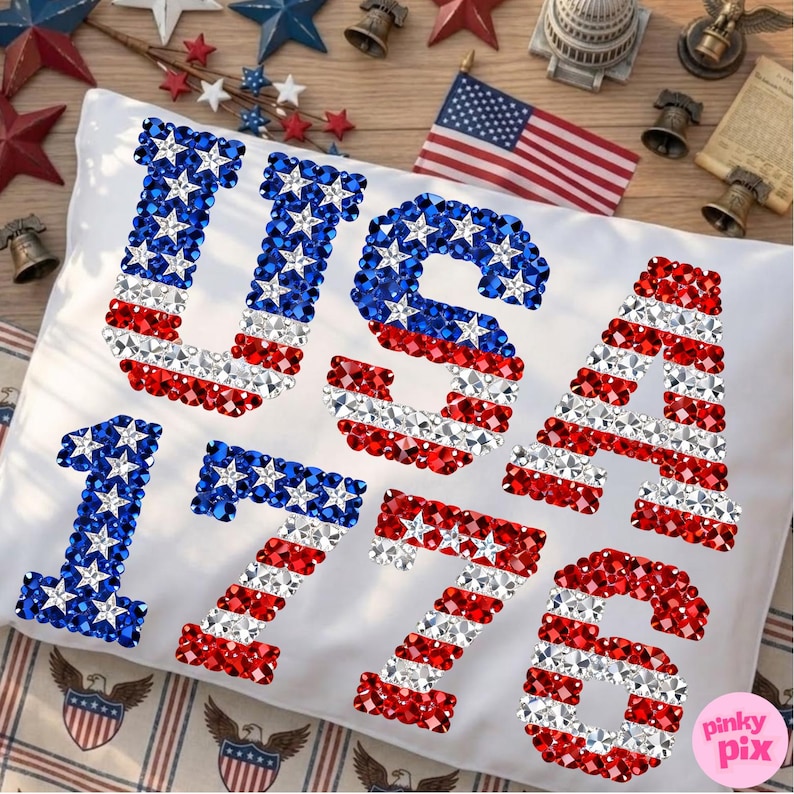 K&ouml;nnte beinhalten: Wei&szlig;es Kissen mit den W&ouml;rtern "USA" und den Zahlen "1776" in rot, wei&szlig; und blau glitzernden Verzierungen, die an die amerikanische Flagge erinnern. Das Kissen liegt auf einer Holzoberfl&auml;che mit patriotischen Dekorationen.