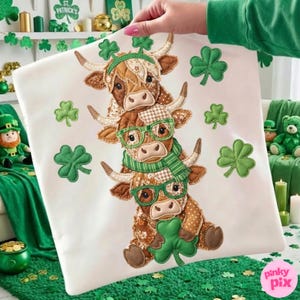 May include: A white pillow featuring a stack of three brown and tan Highland cows, each adorned with green shamrocks and accessories. The cows wear glasses and scarves, with shamrocks scattered around. The pillow is held up by a person.