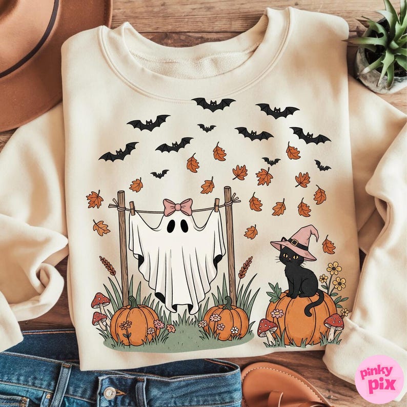 May include: Cream-colored sweatshirt with a Halloween-themed graphic. The design features a ghost with a pink bow, bats, pumpkins, a black cat wearing a witch hat, and falling leaves. The sweatshirt is a cozy fall or Halloween apparel item.