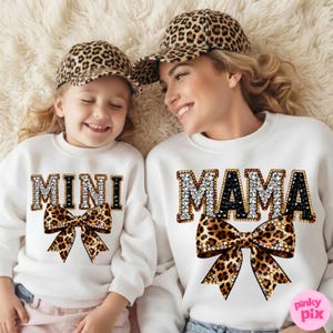 Puede incluir: Dos sudaderas blancas a juego, una con "MAMA" y otra con "MINI" en letras con pedrería, combinadas con lazos con estampado de leopardo. Ambas llevan gorras de béisbol con estampado de leopardo. Imagen de madre e hija.