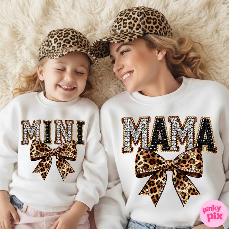 Pu&ograve; includere: Due felpe bianche abbinate, una con "MAMA" e l'altra con "MINI" in lettere con strass, abbinate a fiocchi con stampa leopardata. Entrambe indossano cappellini da baseball con stampa leopardata. L'immagine mostra una madre e una figlia.