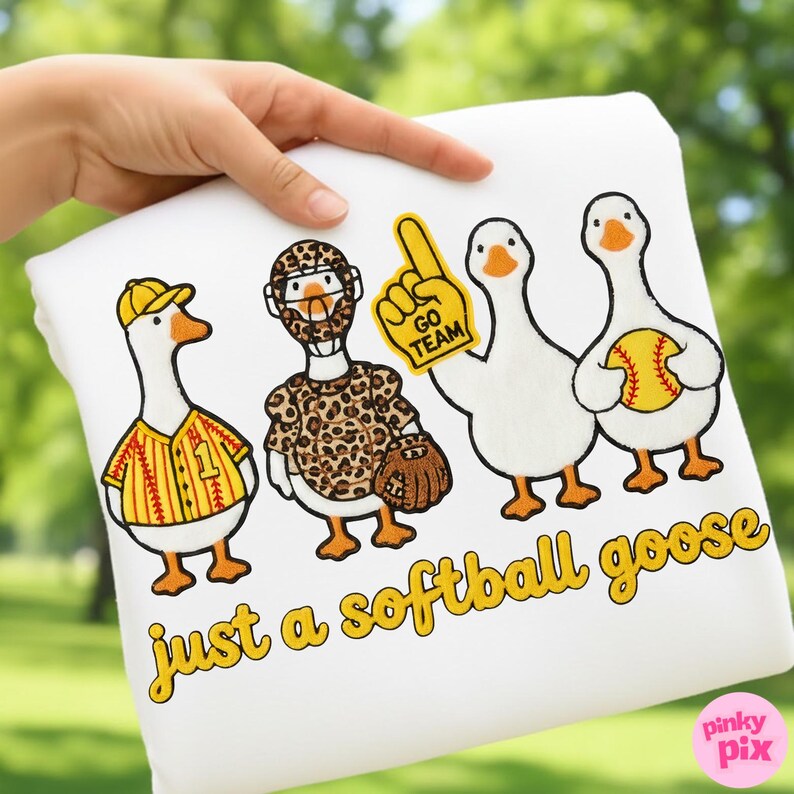 May include: White sweatshirt featuring four cartoon geese dressed in baseball and softball gear. One goose wears a baseball uniform, another a catcher's outfit, and the third holds a foam finger. The text "just a softball goose" is printed in yellow.
