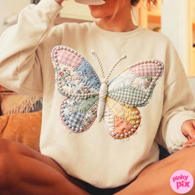 May include: A cream-coloured long-sleeve sweatshirt featuring a large, central butterfly graphic. The butterfly is constructed from various quilted fabric patches in pastel colours, including gingham and floral patterns, outlined with small white beads.