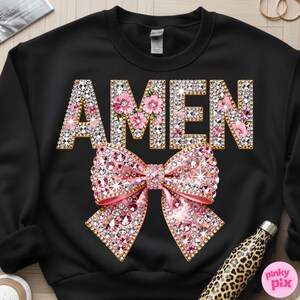 May include: Black sweatshirt featuring the word "AMEN" in gold and rhinestone lettering, with pink floral details. A large pink rhinestone bow is centered below the text. A leopard print water bottle is visible.
