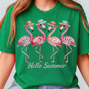 May include: Green t-shirt featuring four pink flamingo illustrations adorned with sparkling embellishments, sunglasses, and hats. The phrase "Hello Summer" is written in a sparkling font below the flamingos. The shirt is a vibrant, summery design.