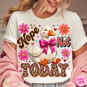May include: White t-shirt with a whimsical design featuring a duck wearing a pink bow, surrounded by flowers and hearts. The shirt has the words "Nope Not Today" in a playful font. The duck has an orange beak and feet.
