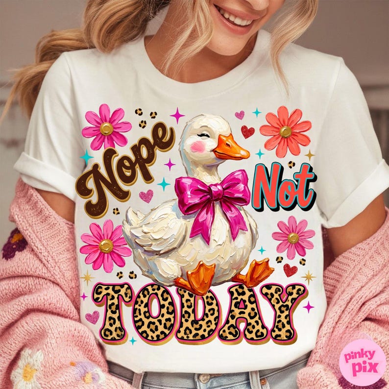 May include: White t-shirt with a whimsical design featuring a duck wearing a pink bow, surrounded by flowers and hearts. The shirt has the words "Nope Not Today" in a playful font. The duck has an orange beak and feet.