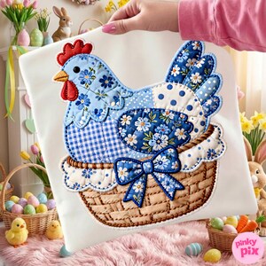May include: A decorative pillow featuring a patchwork hen in a woven basket. The hen is blue with floral and gingham patterns, a red comb, and a yellow beak. A blue bow adorns the basket. The pillow is held up by a person.