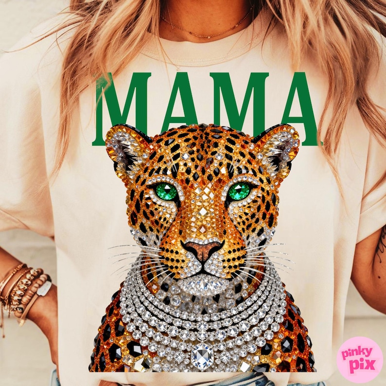May include: A cream-coloured t-shirt with the word "MAMA" in green block letters. The shirt features a detailed leopard design with jewel-toned embellishments, including a jewelled necklace and emerald green eyes.