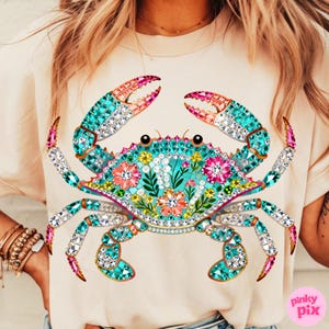 May include: A light beige t-shirt featuring a detailed, jewel-toned crab design. The crab has turquoise, pink, and clear crystal accents, with floral details on its body. The design is centered on the shirt.