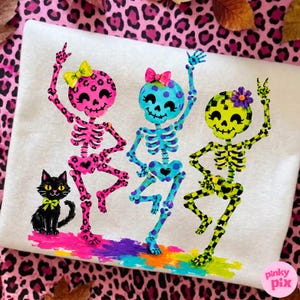 May include: A white t-shirt featuring a graphic of three dancing skeletons and a black cat. The skeletons are pink with leopard print, blue with polka dots, and lime green with a checkerboard pattern, each wearing a colorful bow or flower.