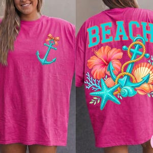 May include: A magenta t-shirt featuring a turquoise anchor design on the front and the word "BEACH" on the back. The back design includes a turquoise anchor, starfish, and seashell graphics with a hibiscus flower.