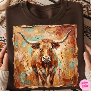 May include: Brown sweatshirt featuring a textured painting of a longhorn steer. The artwork uses a palette of warm browns, oranges, and teal, with visible brushstrokes. The steer faces forward, showcasing its large horns. The sweatshirt is held by a person.
