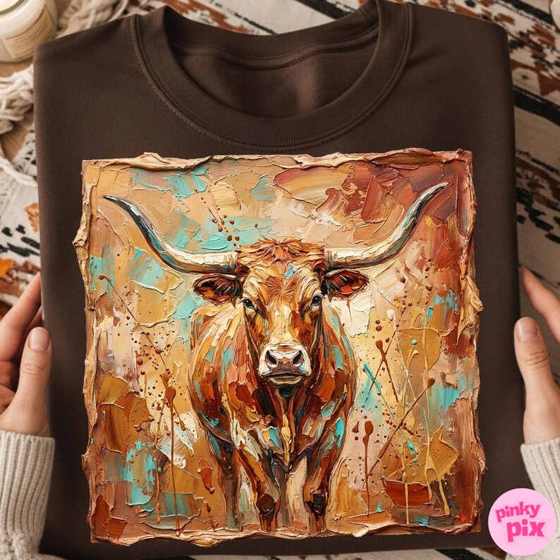May include: Brown sweatshirt featuring a textured painting of a longhorn steer. The artwork uses a palette of warm browns, oranges, and teal, with visible brushstrokes. The steer faces forward, showcasing its large horns. The sweatshirt is held by a person.
