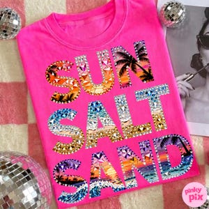 May include: A bright pink t-shirt with the words "SUN SALT SAND" in a jeweled design. The letters feature sunset and beach scenes with palm trees and waves. The shirt is a crew neck style.