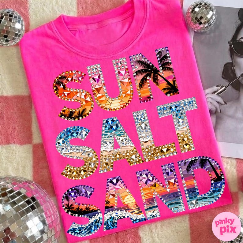 May include: A bright pink t-shirt with the words "SUN SALT SAND" in a jeweled design. The letters feature sunset and beach scenes with palm trees and waves. The shirt is a crew neck style.