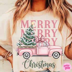 May include: A light beige t-shirt featuring a pink vintage truck carrying a Christmas tree with ornaments and a large bow. The words "MERRY CHRISTMAS" are printed in pink and green.