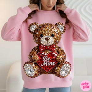 May include: Pink sweatshirt featuring a large, jeweled teddy bear design. The bear has a red bow tie and a heart-shaped chest with the words "Be Mine". The bear is made of brown and clear crystals.