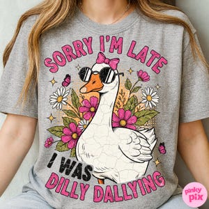 K&ouml;nnte beinhalten: Graues T-Shirt mit einem Motiv einer wei&szlig;en Gans mit Sonnenbrille und rosa Schleife, umgeben von Blumen und Schmetterlingen. Der Text lautet "SORRY I'M LATE I WAS DILLY DALLYING" in rosa und schwarzer Schrift.