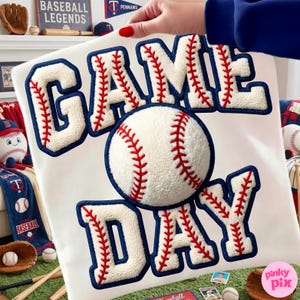 May include: A white baseball-themed pillow with the words "GAME DAY" in large, textured letters. The letters are white with red stitching and blue outlines. A baseball is in the center. Baseball-themed items are in the background.
