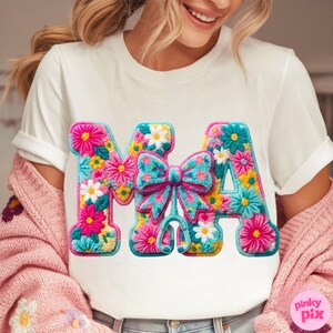 May include: White t-shirt with a colorful floral design spelling out "MA" with a bow and stethoscope. The letters are filled with pink, yellow, blue, and white flowers. The bow is turquoise with a pink outline.