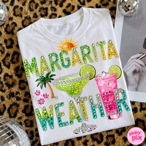 Peut inclure: T-shirt blanc avec l'inscription "MARGARITA WEATHER" en strass verts, jaunes et bleus scintillants. Le motif comprend un verre &agrave; margarita, un cocktail rose, un soleil, un palmier et des tranches de citron vert.