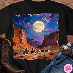 May include: Black t-shirt featuring a vibrant painting of cowboys riding horses through a canyon under a full moon. The artwork showcases a night sky with purple clouds and orange rock formations. A cowboy boot and a belt buckle are visible.
