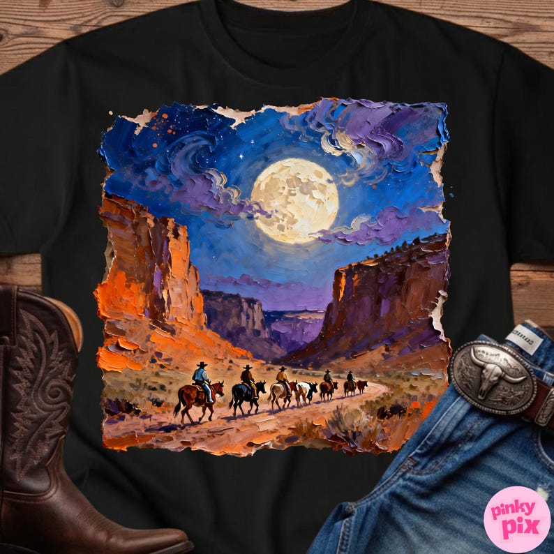 May include: Black t-shirt featuring a vibrant painting of cowboys riding horses through a canyon under a full moon. The artwork showcases a night sky with purple clouds and orange rock formations. A cowboy boot and a belt buckle are visible.