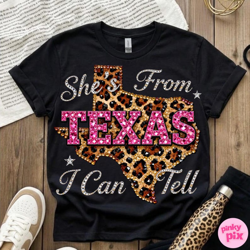 May include: Black t-shirt with the text "She's From Texas I Can Tell". The word "TEXAS" is in pink, and the outline of Texas is in a leopard print design. The text is embellished with rhinestones.