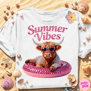 May include: White t-shirt with the text "Summer Vibes" in pink rhinestone lettering. The shirt features a graphic of a Highland cow wearing sunglasses and a rhinestone-studded floatie. The cow is surrounded by seashells and pearls.
