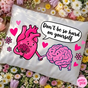 May include: White pillow with a pink heart and brain graphic holding hands. The heart holds flowers. Text in a speech bubble reads "Don't be so hard on yourself." Pink hearts and flowers decorate the pillow.