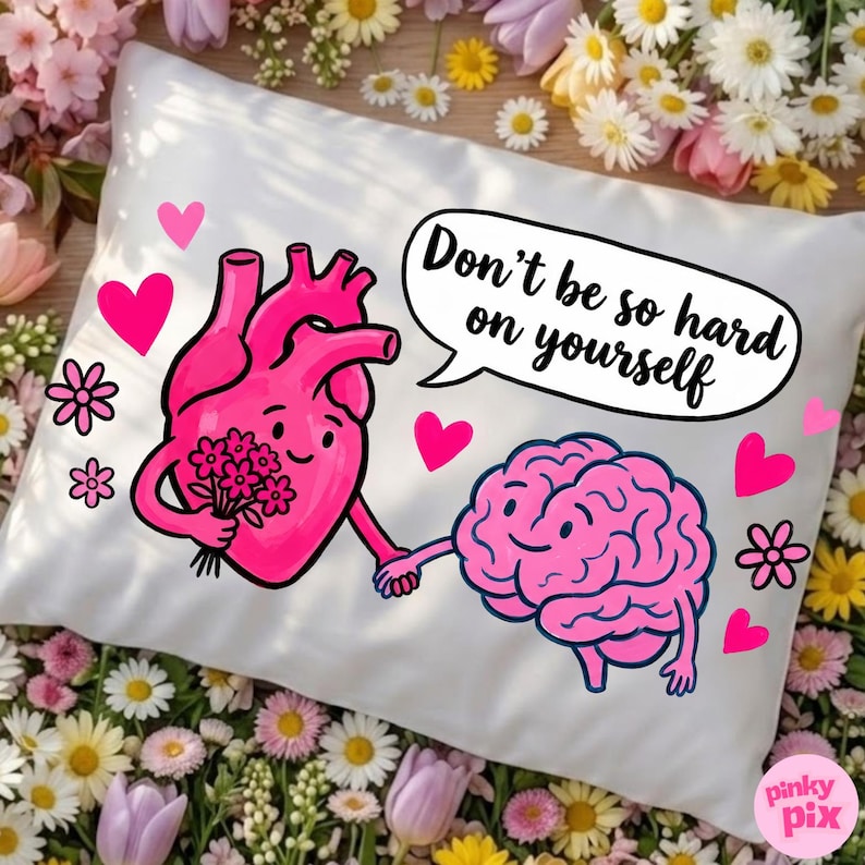 May include: White pillow with a pink heart and brain graphic holding hands. The heart holds flowers. Text in a speech bubble reads "Don't be so hard on yourself." Pink hearts and flowers decorate the pillow.