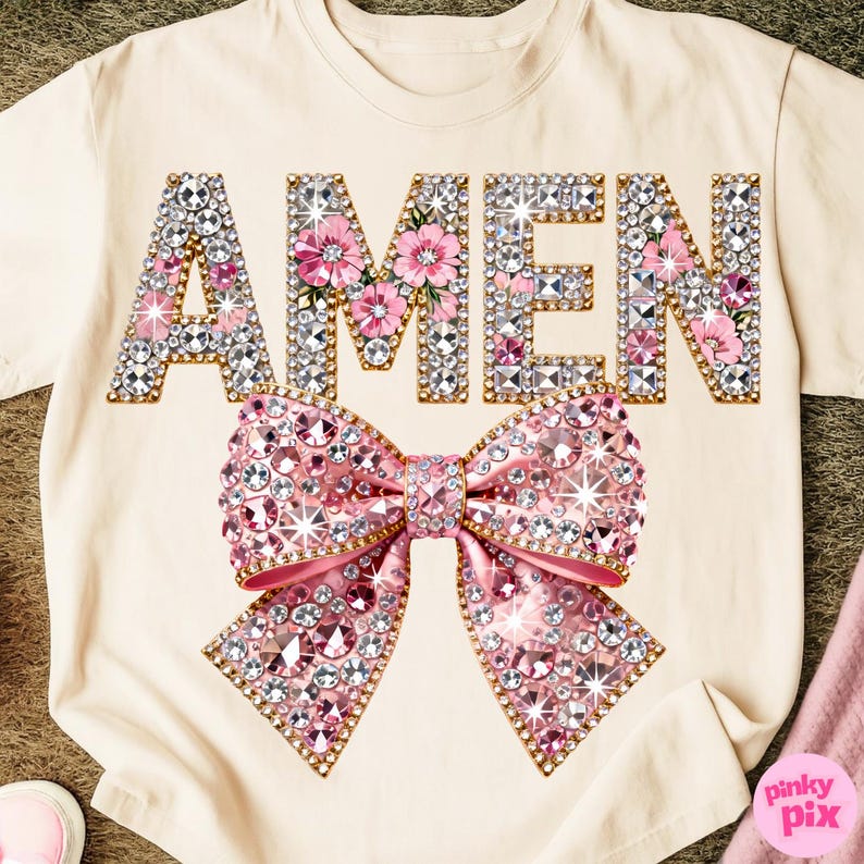 May include: A cream-coloured t-shirt featuring the word "AMEN" in a jewelled design, with pink floral accents. A large pink jewelled bow is centered below the text. The shirt has a casual, feminine style.