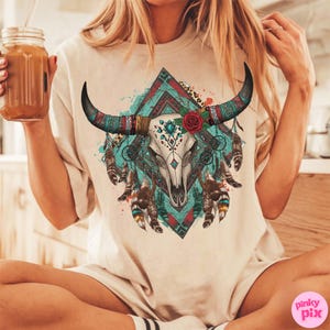 May include: A cream-coloured t-shirt with a detailed graphic of a bull skull with horns, decorated with a red rose, turquoise accents, and feathers. The design is set against a teal and red geometric background, creating a bohemian aesthetic.